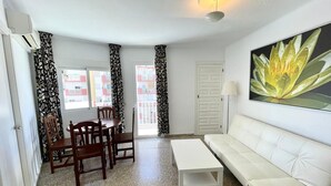 A living room with a white sofa, a wooden dining table with chairs, a white coffee table, patterned curtains, a door, and a wall-mounted lamp.