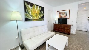 A living room with a white sofa, a white coffee table, a floor lamp, a TV on a wooden cabinet, and a painting of a flower on the wall.