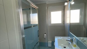 Un baño moderno con cabina de ducha de vidrio, un lavabo blanco con espejo y una ventana que proporciona luz natural.
