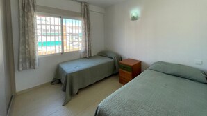 Una habitación con dos camas individuales, una mesita de noche de madera, una ventana con cortinas y una lámpara fijada a la pared.