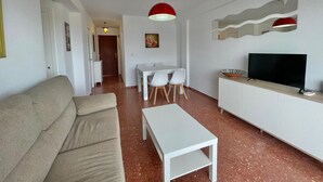 Un saló amb un sofà, una taula de cafè blanca, una televisió sobre un moble, una zona de menjador amb taula i cadires, i un passadís que condueix a una porta.