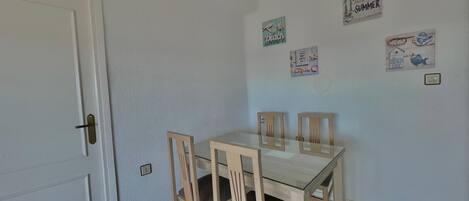 A dining area with a glass-top table and four wooden chairs. There are wall decorations including a 'Hello Summer' sign and a 'Hello Days' sign.