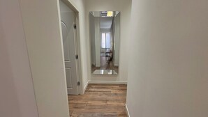 Un pasillo con suelo de madera y una pared espejada que refleja una habitación con ventana y cortinas.
