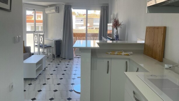 A modern kitchen with white cabinets, a dining area with a white table and chairs, and a view of buildings through large windows. - TERRASOL PLAZAMAR 6-2 (Torre del Mar)