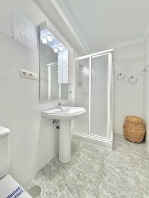 A bathroom with a pedestal sink, a mirror with lights, a shower enclosure, and a wicker basket.