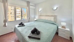 Ein Schlafzimmer mit Bett, Nachttisch, einem Fenster mit Vorhängen und Blick auf Gebäude.