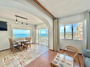 A spacious room with a dining area, a balcony with a view of the sea, and a large window overlooking a cityscape.