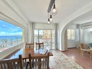 A room with a large window overlooking a beach, a balcony with a view of the sea, a dining table with chairs, and a patterned rug.