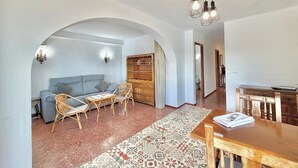 Ein Wohnzimmer mit Sofa, Sesseln, Couchtisch, Teppich mit Muster, Holzschrank und Esstisch mit Stühlen.