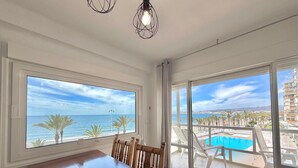Ein Zimmer mit großem Fenster zum Strand, einem Esstisch sowie Blick auf Pool und Gebäude.