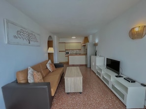 Een woonkamer met een bank, salontafel, televisie en een keuken op de achtergrond.
