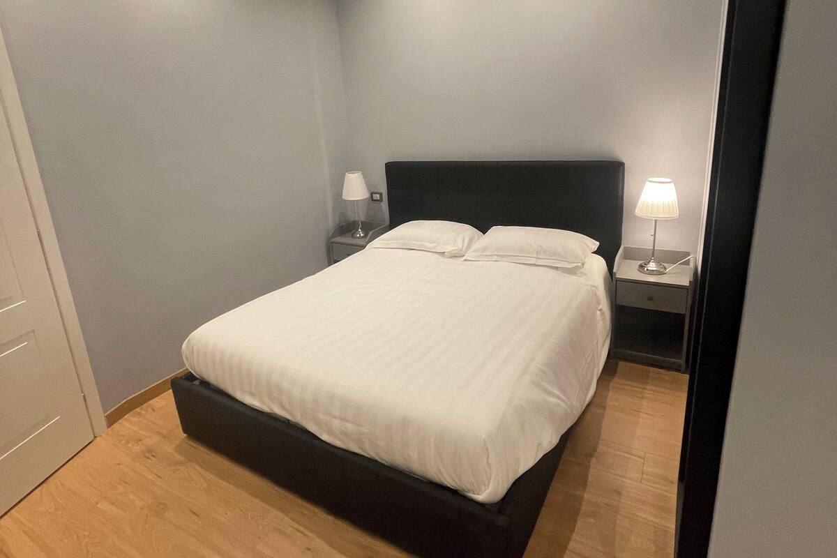 A bedroom with a bed, two bedside tables with lamps, a wooden floor, and a ceiling with recessed lighting.