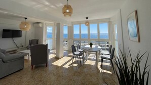Un salón amplio con una mesa de comedor y sillas, un sofá gris y un televisor. Ventanas grandes ofrecen vistas al mar.