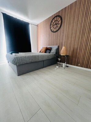 A modern bedroom with a bed, a wall clock, and a small table with a lamp. - Nest And Limited (London)