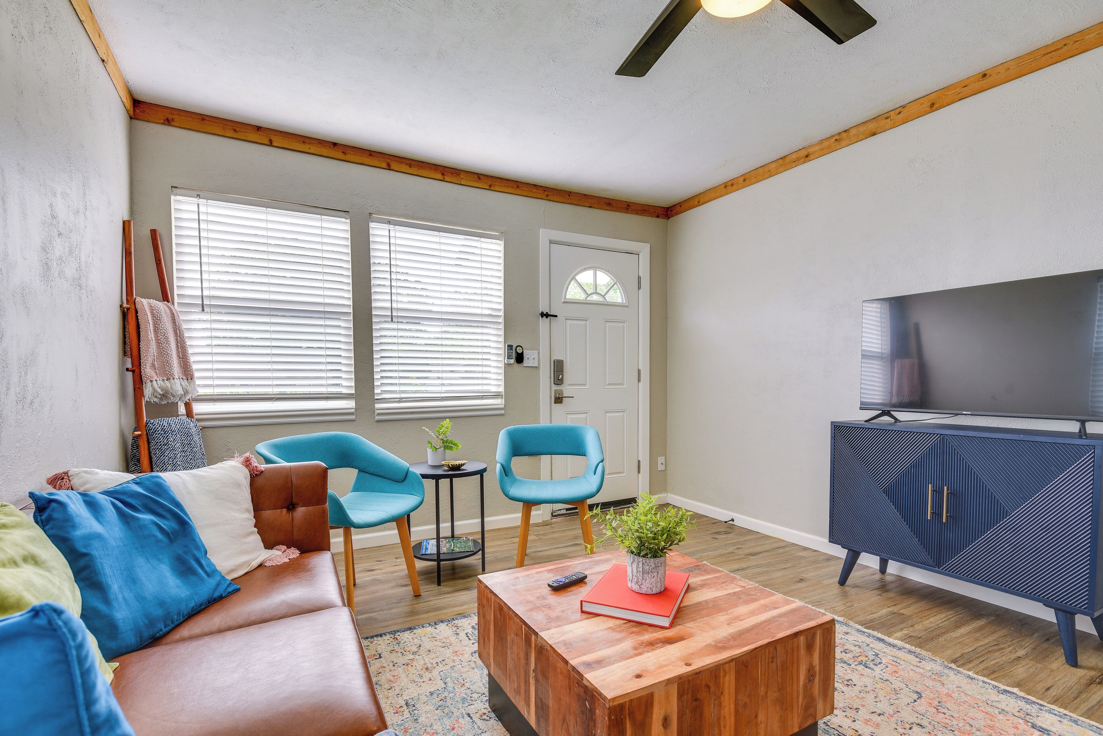 Una sala de estar con un sofá de cuero marrón, dos sillones azules, una mesa de centro de madera con un libro y una planta, un mueble azul con un televisor, y una puerta con ventanal.