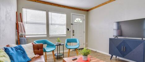 Una sala de estar con un sofá de cuero marrón, dos sillones azules, una mesa de centro de madera con un libro y una planta, un mueble azul con un televisor, y una puerta con ventanal.