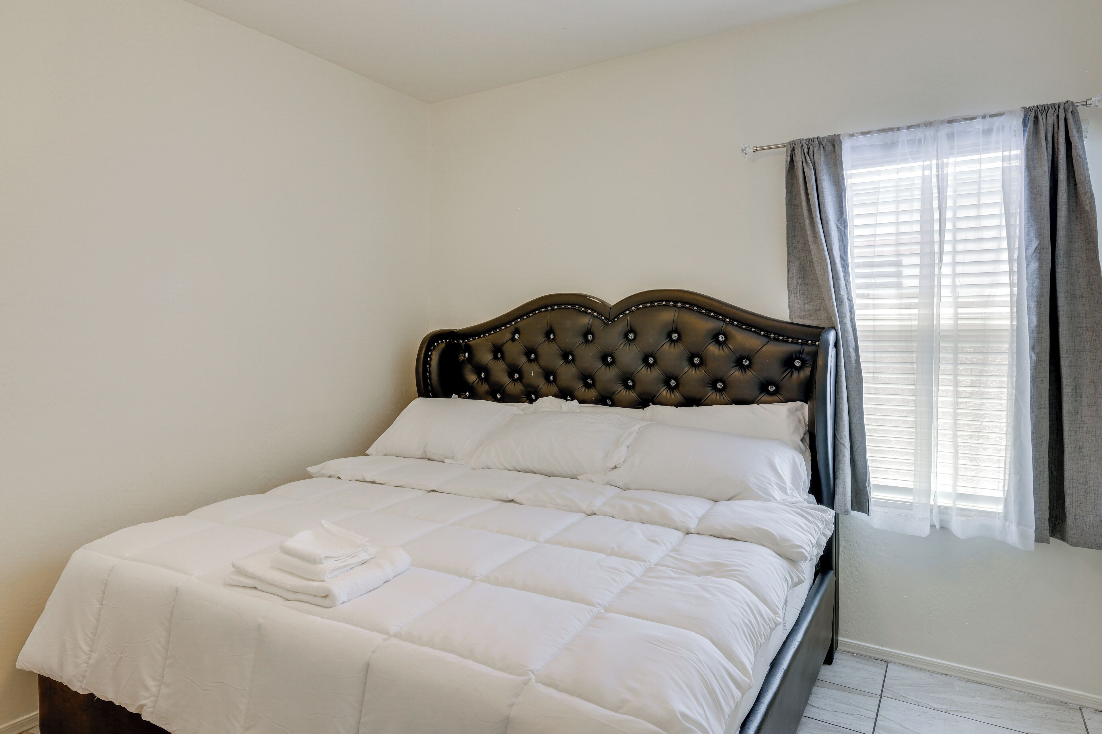 A neatly made bed with white bedding and a dark headboard in a room with white walls and a window with curtains.