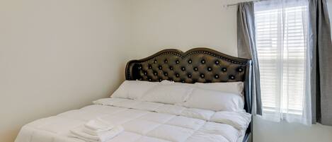 A neatly made bed with white bedding and a dark headboard in a room with white walls and a window with curtains.