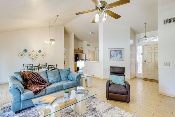A living room with a blue sofa, a glass coffee table, a brown leather chair, and a ceiling fan. - Southwest Getaway w/ Fenced Yard in Chandler! (Chandler)