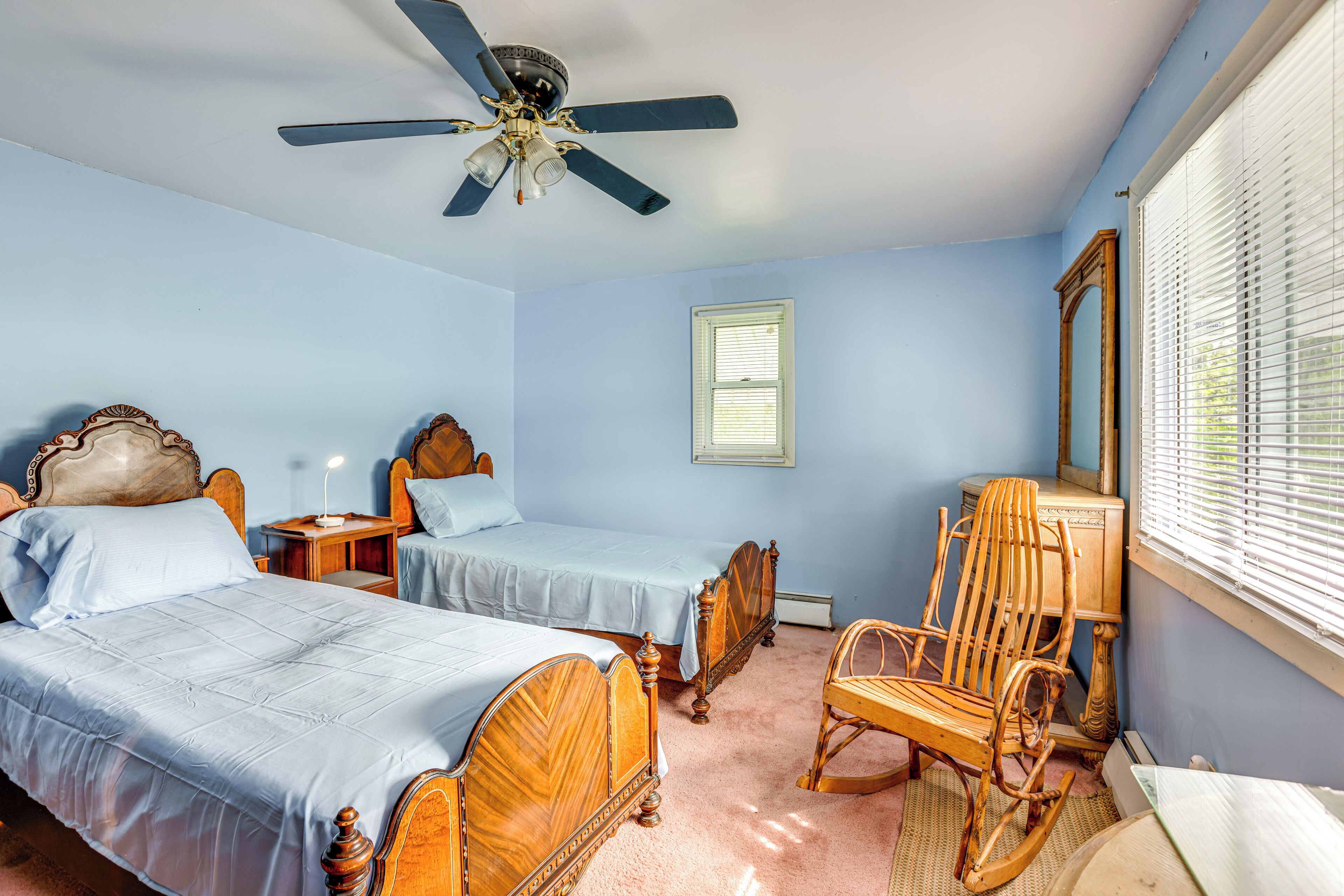 A bedroom with two wooden beds, a ceiling fan, a rocking chair, and a window with blinds.