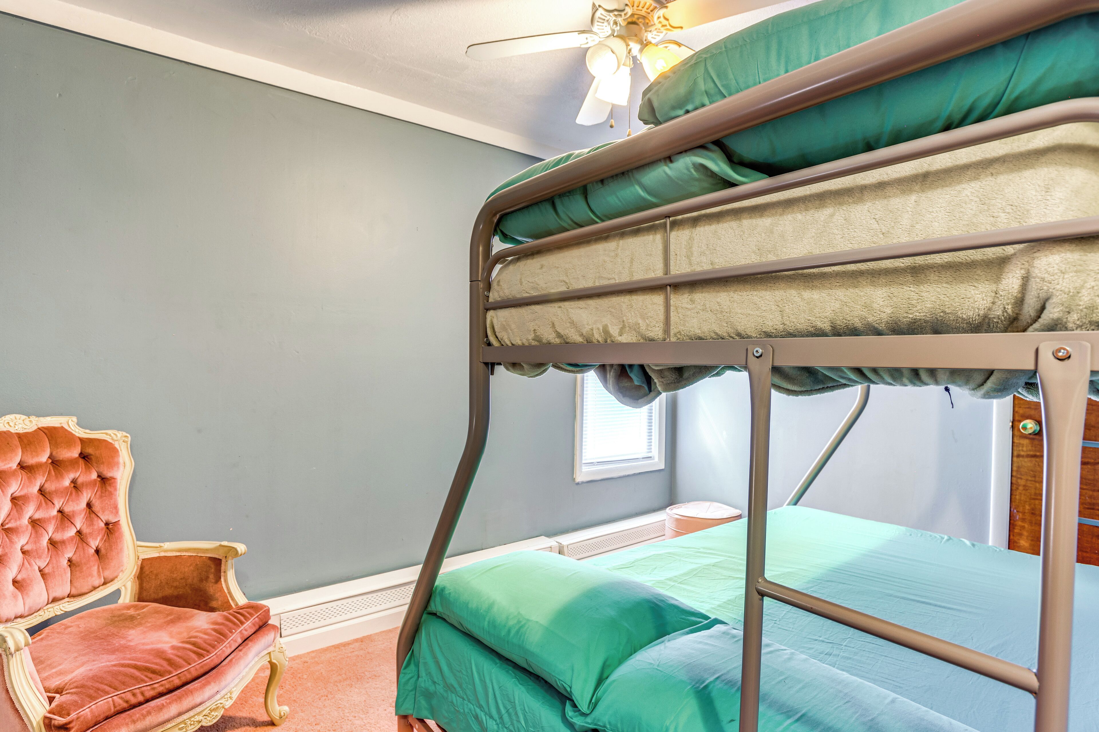 Uma cama beliche com roupa de cama verde, uma cadeira antiga e um ventilador de teto num quarto com paredes azul claro.
