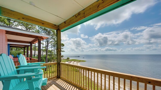 Een houten terras met turquoise Adirondack-stoelen, uitzicht op het strand en een pier in de verte.