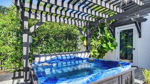 Un bain à remous installé sous une pergola, avec une porte blanche et une végétation luxuriante en arrière-plan.