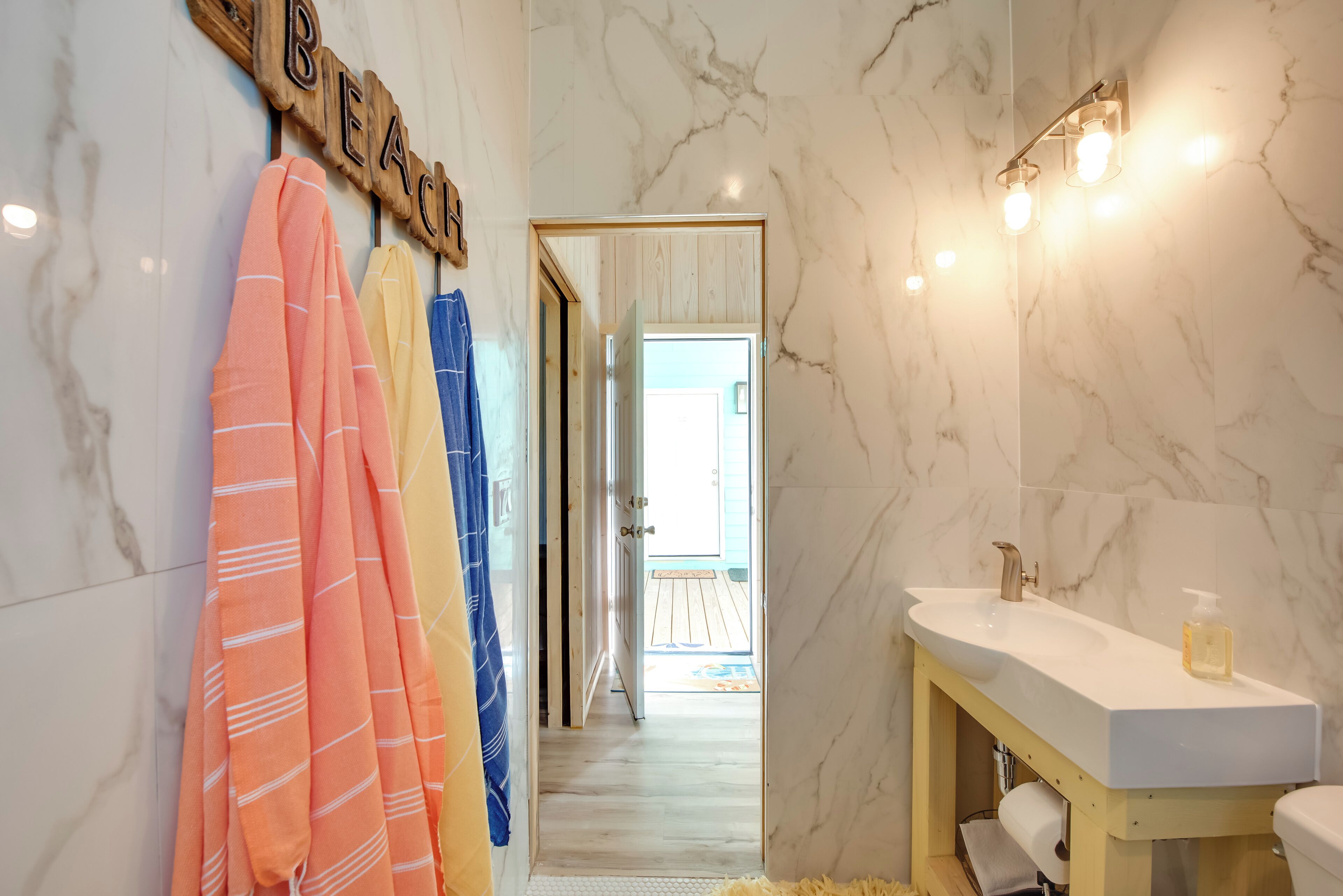A bathroom with a marble wall, a wooden towel rack with towels, a sink with a soap dispenser, and a mirror with lights.
