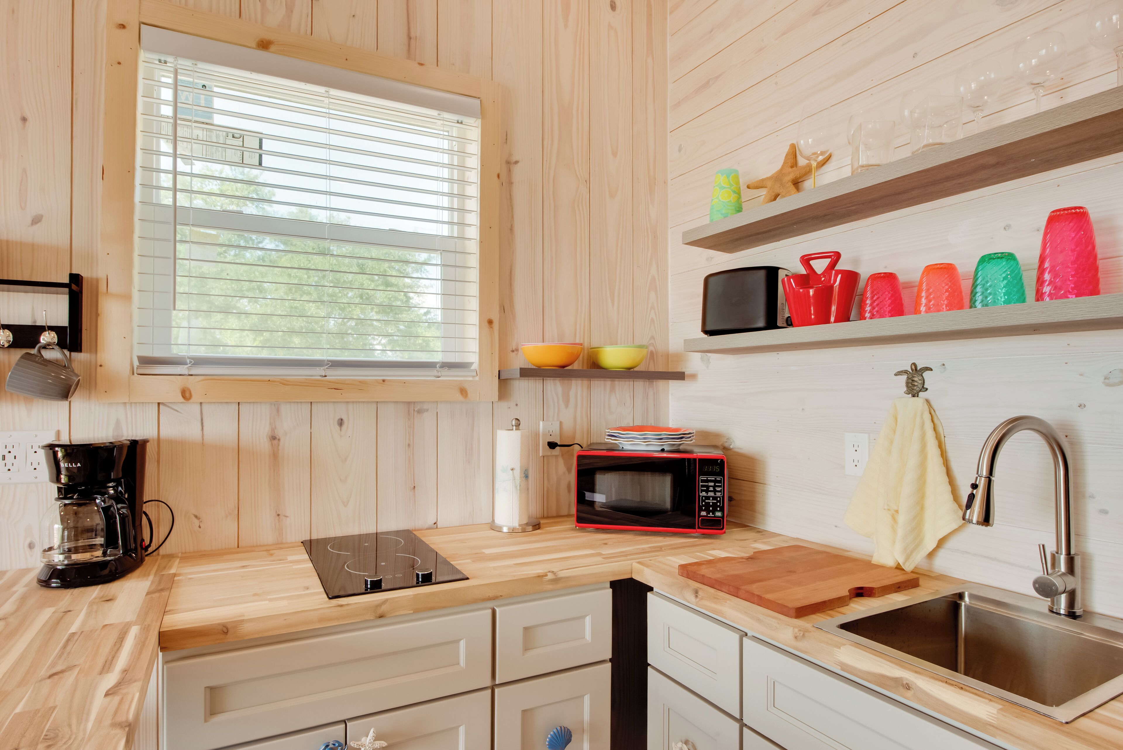 A modern kitchen with wooden cabinetry, a black coffee maker, a microwave, a cutting board, and a stainless steel sink.