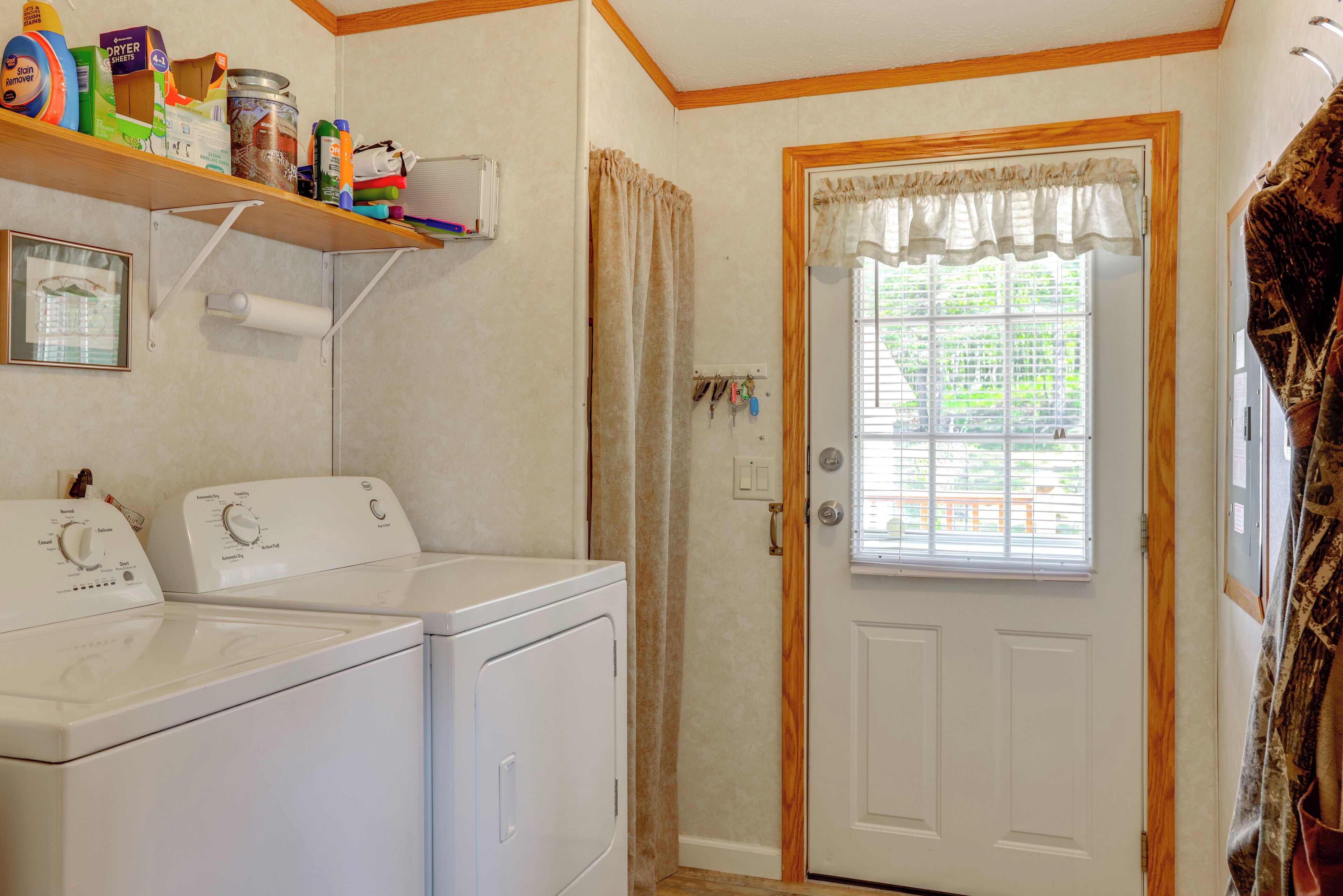 A laundry room with two white washers and dryers, a shelf with cleaning supplies, a door with a window, and a curtain.