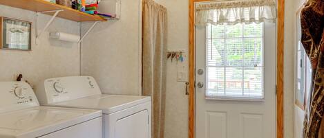 A laundry room with two white washers and dryers, a shelf with cleaning supplies, a door with a window, and a curtain.