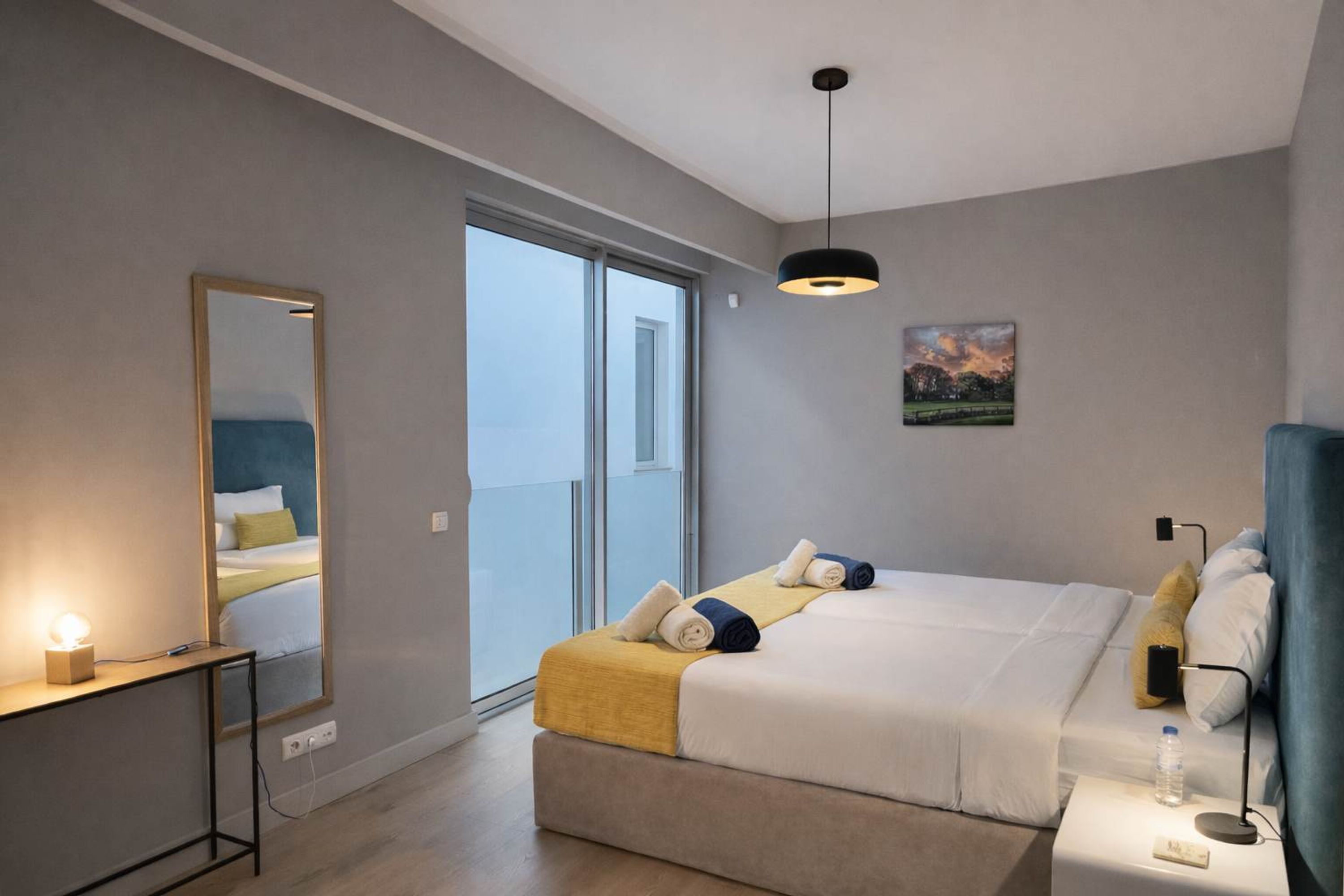 A modern hotel room with a large bed, a bedside table with a lamp, a mirror, and a view of the outdoors.
