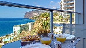 Una mesa de desayuno con croissants, zumo de naranja y tazas de café, con vistas a la costa, edificios y una montaña.