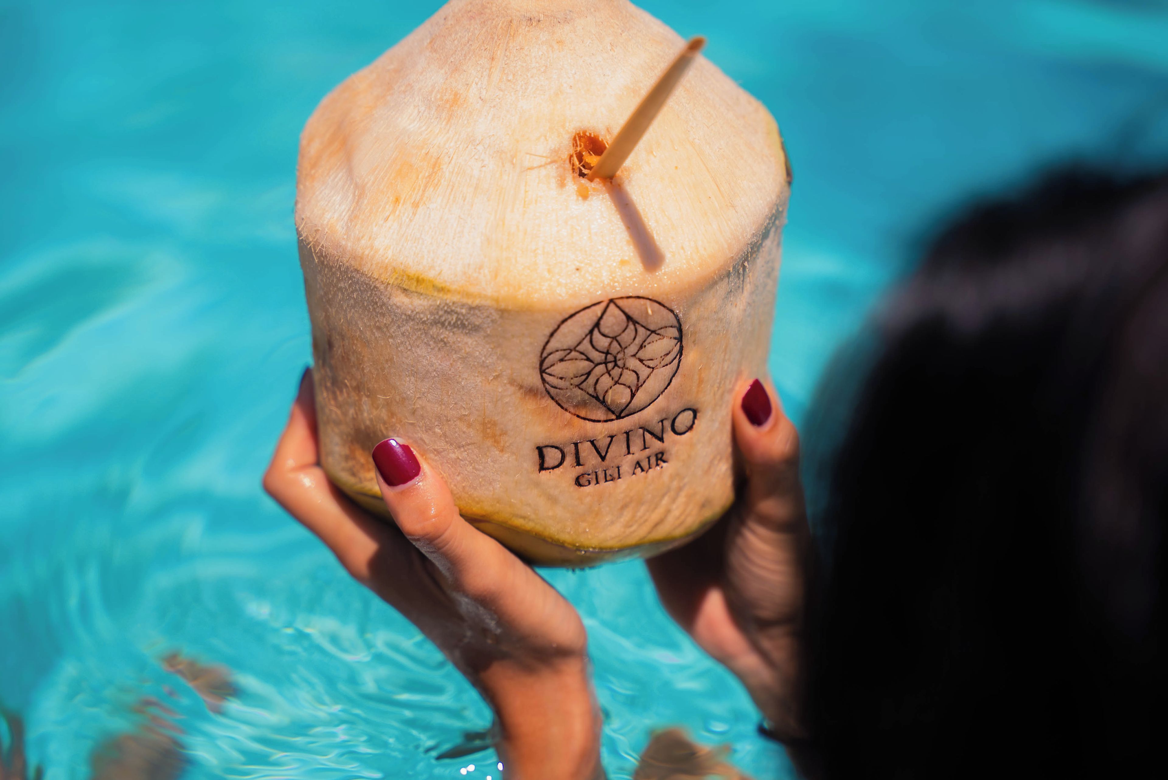 Una persona sostiene un coco con una pajita, con la etiqueta 'DIVINO GILI AIR' en una piscina.