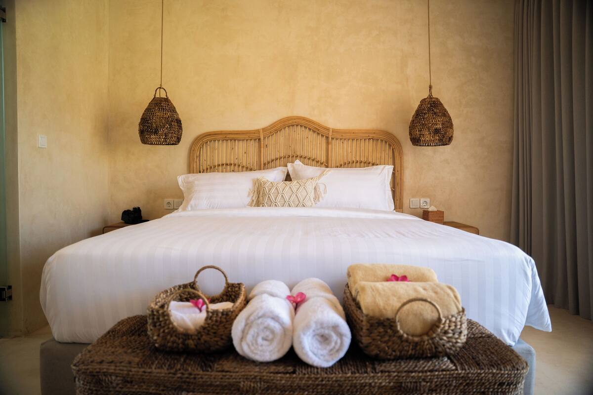 A neatly made bed with white linens, two hanging woven lamps, and a basket of rolled towels on a bedside table.