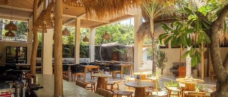 A restaurant with wooden tables and chairs, thatched roof, and tropical plants.