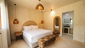 A bedroom with a large bed, bedside tables, a bench, a mirror, and a bathroom with a sink and mirror. - Divino Gili Air (Gili Air)