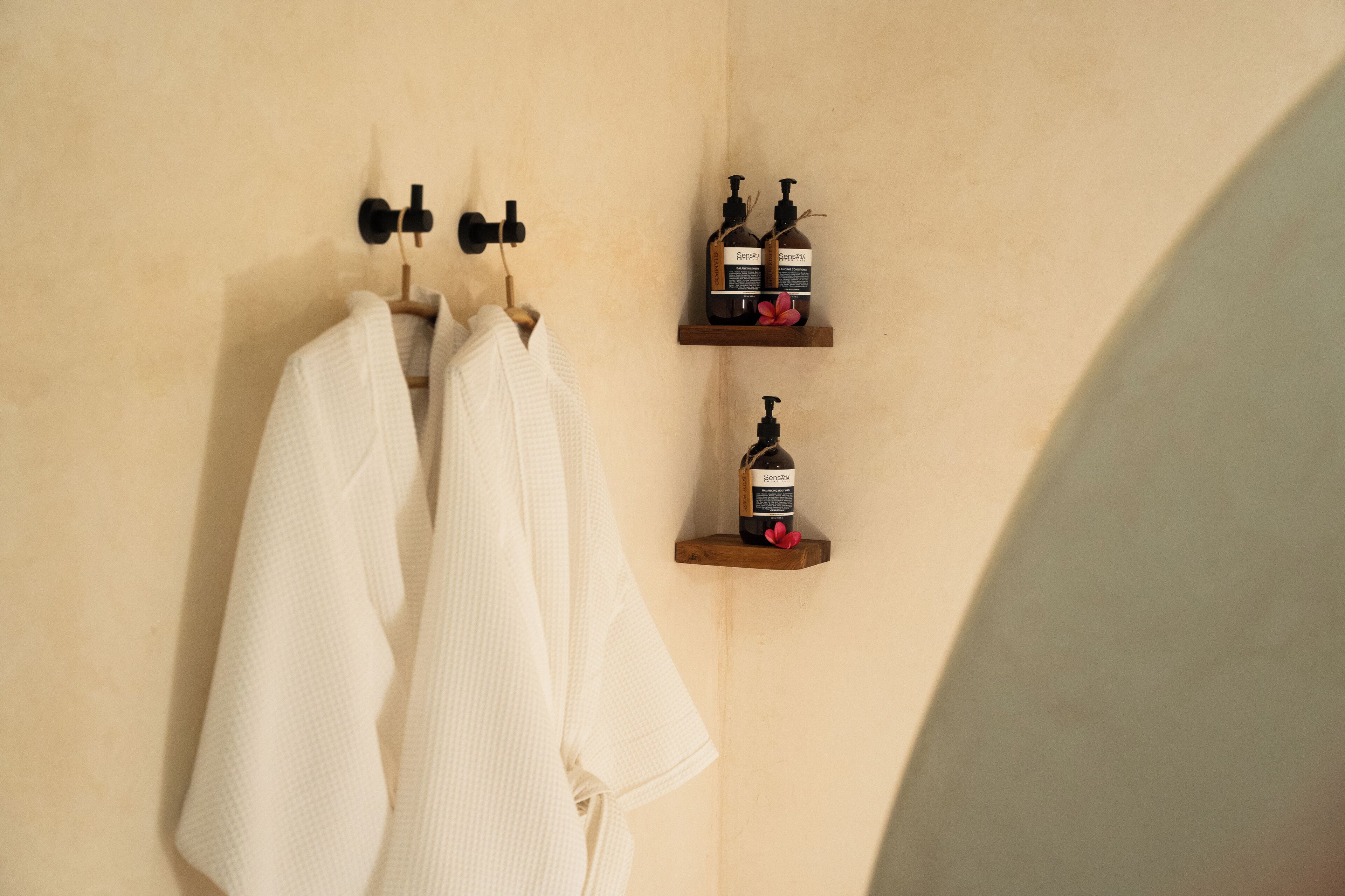 Un baño con dos toallas blancas colgadas en un perchero, un estante de madera con dos botellas de Sensatia para el cuerpo y una sola botella de loción Sensatia.