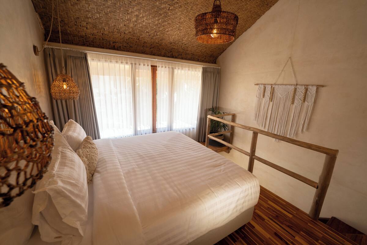 A bedroom with a large bed, a woven headboard, a hanging light fixture, and a view of the outdoors through sheer curtains.