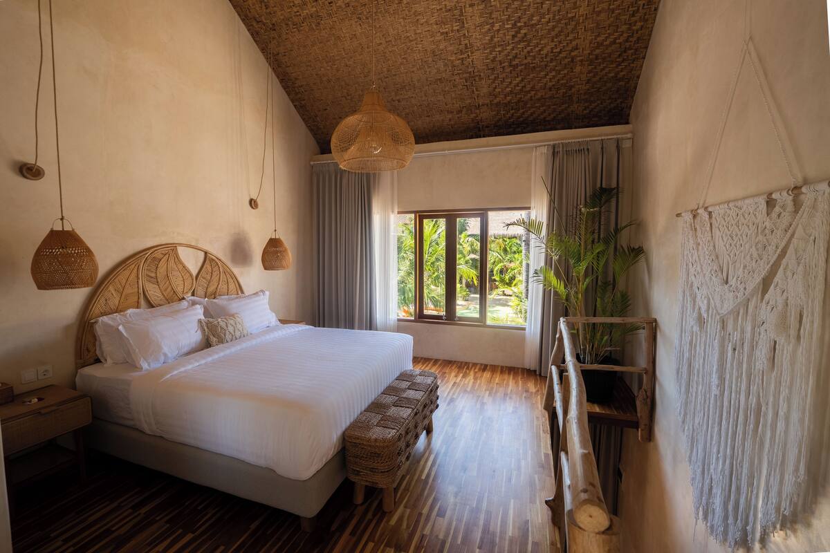 A bedroom with a large bed, wooden floor, woven headboard, hanging wicker lamps, and a view of greenery outside.