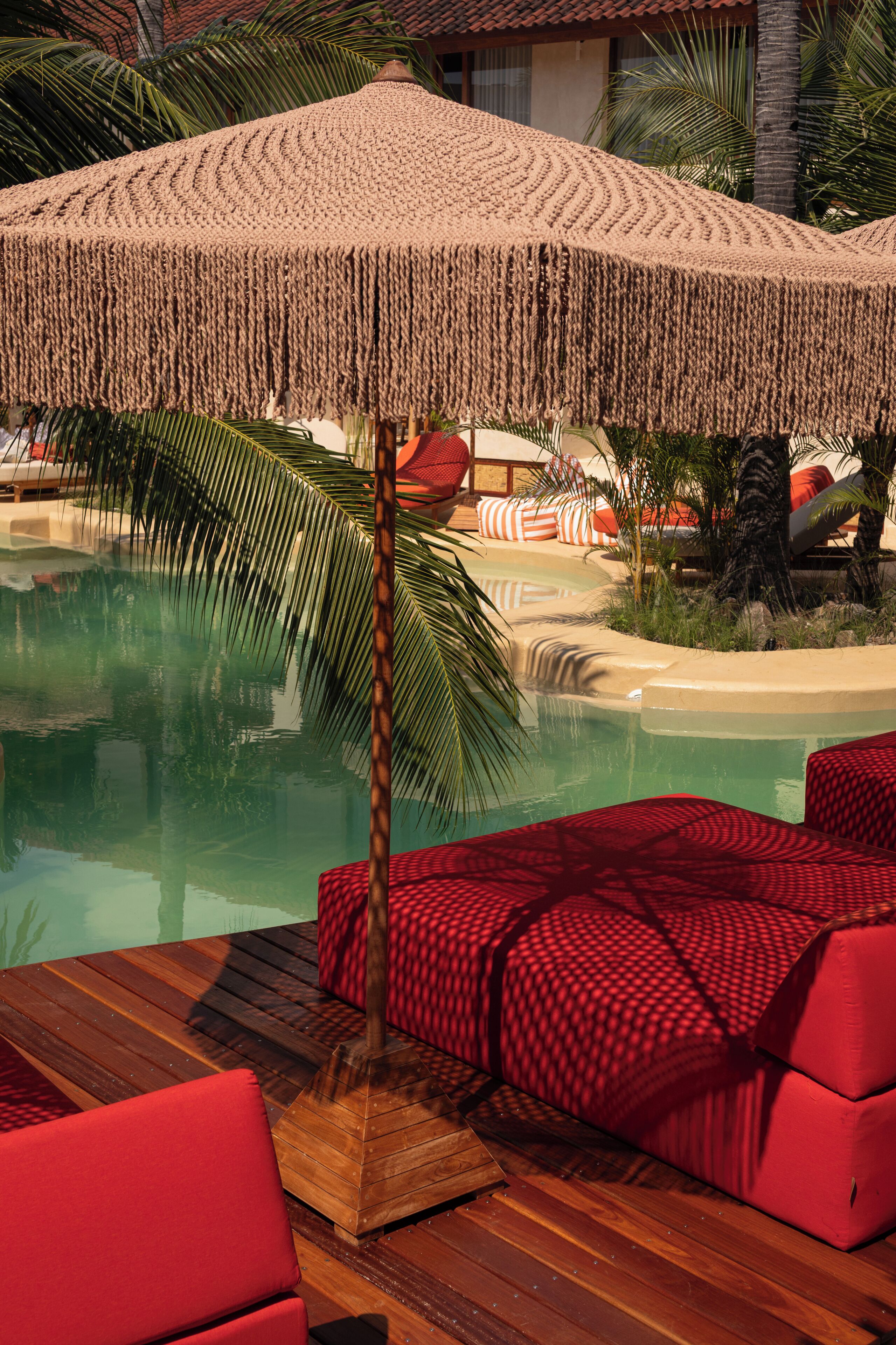 A poolside area with a wooden deck, red cushioned seating, and a large woven umbrella.