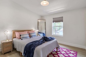 A bedroom with a bed, bedside table, mirror, window, and a pink patterned rug. - The Jasmine Loft (Tybee Island)