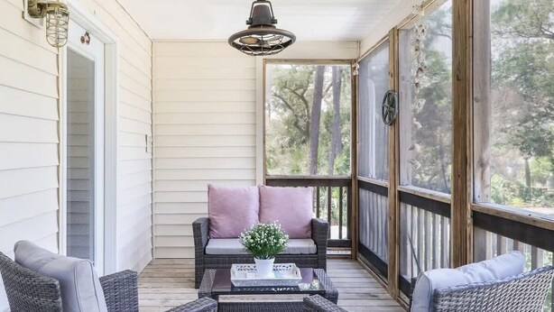 A covered porch with wicker furniture, a coffee table with a floral centerpiece, and a view of trees through the glass door. - The Jasmine Loft (Tybee Island)
