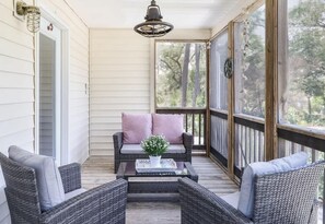 A covered porch with wicker furniture, a coffee table with a floral centerpiece, and a view of trees through the glass door. - The Jasmine Loft (Tybee Island)