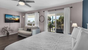 Un dormitorio con una cama grande, un ventilador de techo, televisión, balcón con cortinas y vistas a palmeras.