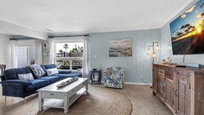 Una sala de estar con un sofá azul, una mesa de centro blanca, un sillón estampado, un mueble de madera y un cuadro grande de un atardecer.