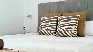 A neatly made bed with a grey headboard, two patterned pillows, and a small woven stool with glasses on it. - Canaryislandshost l Amapola (Las Palmas)