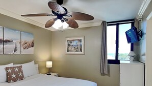 A bedroom with a bed, a ceiling fan, a nightstand with a lamp, a framed picture, a window with curtains, and a TV mounted on the wall. - Sunbird Beach Resort #902 - Oceanfront Escape | Gulf Front (Panama City Beach)
