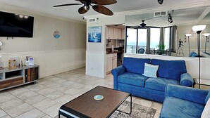 A living room with a blue sofa, a coffee table, a TV, and a ceiling fan. - Sunbird Beach Resort #902 - Oceanfront Escape | Gulf Front (Panama City Beach)