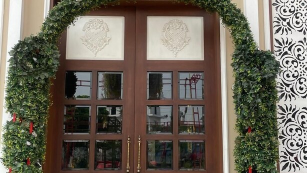 Uma porta dupla de madeira com painéis de vidro, emoldurada por painéis decorativos e uma coroa de flores verdes.